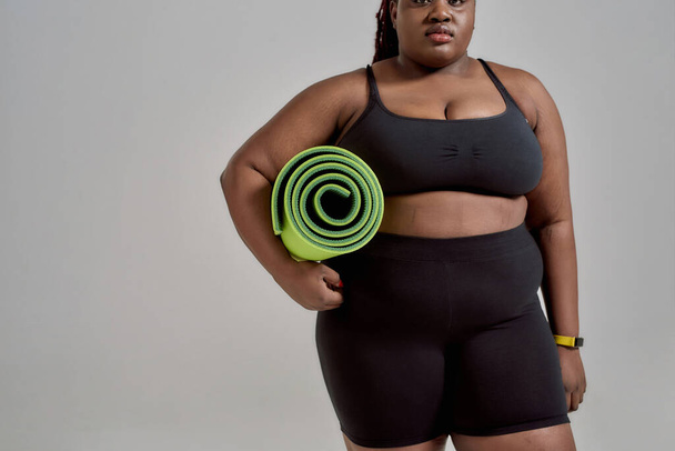 Yoga time. Cropped shot of plump, plus size african american woman in sportswear holding green yoga mat for fitness, posing in studio over grey background - Foto, imagen