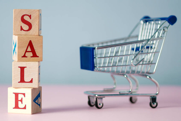 Red word SALE laid out of wooden cubes with an empty supermarket food cart on grey and pink background with space for text. Concept of selling products in supermarket or online store on Black Friday - 写真・画像