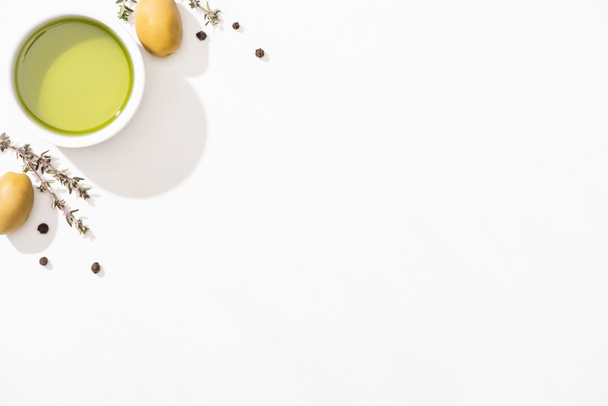 вид на оливковое масло в миске рядом с зелеными оливками, травами и черным перцем на белом фоне
 - Фото, изображение