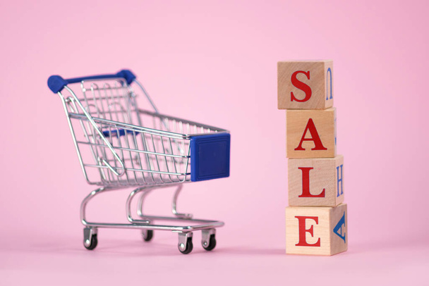 Red word SALE laid out of wooden cubes with an empty supermarket cart for products on pink background with space for text. Concept of selling products in supermarket or online store on Black Friday - Φωτογραφία, εικόνα