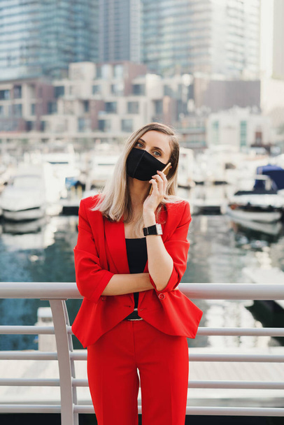 Mladá podnikatelka v červeném obleku a ochranné masky stojí na promenádě ve městě a telefonuje přes chytrý telefon - Fotografie, Obrázek