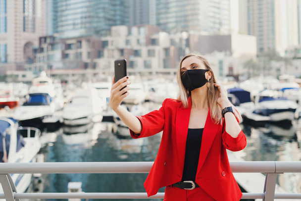 Mujer rubia joven en traje rojo y con mascarilla protectora tomando una foto selfie cerca de yates de lujo y rascacielos. Nuevo concepto normal
 - Foto, imagen
