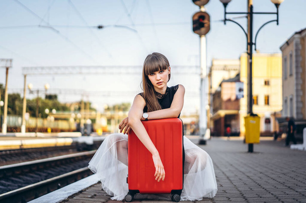 Female brunette traveler with red suitcase in white skirt waiting for a train on raiway station - Φωτογραφία, εικόνα