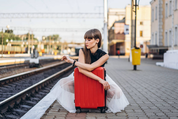 Female brunette traveler with red suitcase in white skirt waiting for a train on raiway station and checking time on her wristwatch - Foto, afbeelding