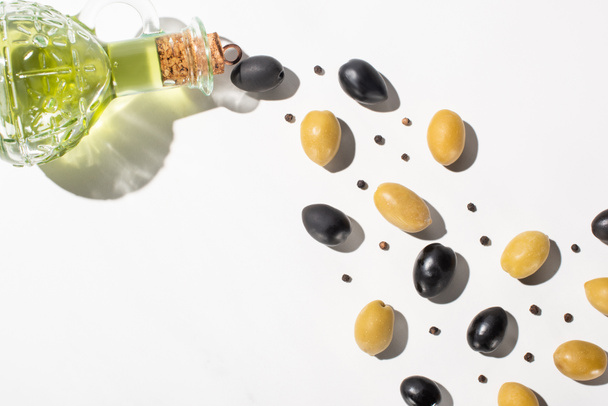 Ansicht von Olivenöl in der Flasche in der Nähe von grünen und schwarzen Oliven und schwarzem Pfeffer auf weißem Hintergrund mit Schatten - Foto, Bild