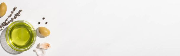 κορυφαία άποψη του ελαιολάδου σε μπολ κοντά σε πράσινες ελιές, σκόρδο, βότανο και μαύρο πιπέρι σε λευκό φόντο, πανοραμικός προσανατολισμός  - Φωτογραφία, εικόνα