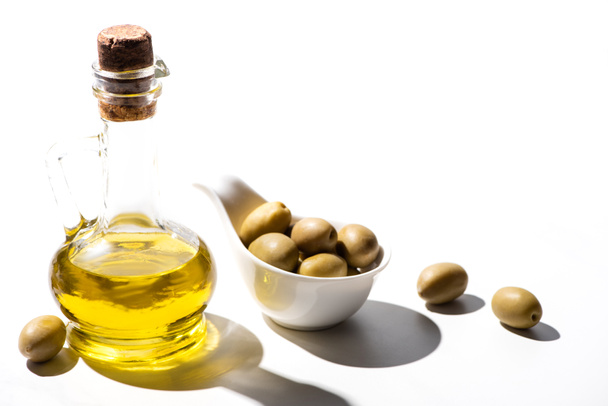 olijfolie in pot bij groene olijven in schaal op witte achtergrond - Foto, afbeelding