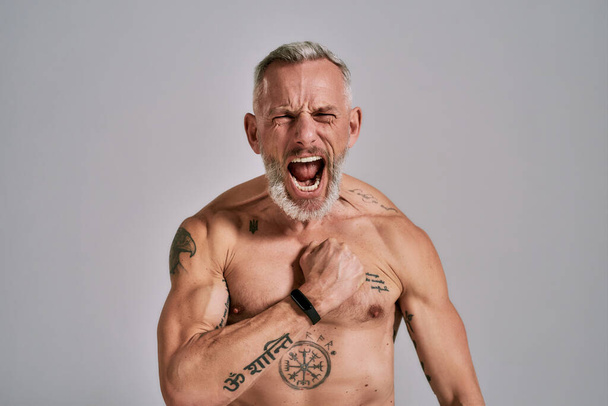 Inner power. Close up of half naked middle aged muscular man shouting at camera, beating his chest while posing in studio over grey background - Photo, image