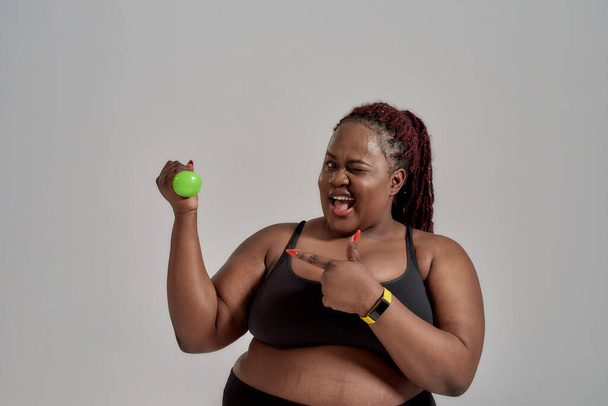 The Choice is Clear. Cheerful plump, plus size african american woman in sportswear pointing at arm holding green dumbbell, while exercising in studio over grey background - Photo, image