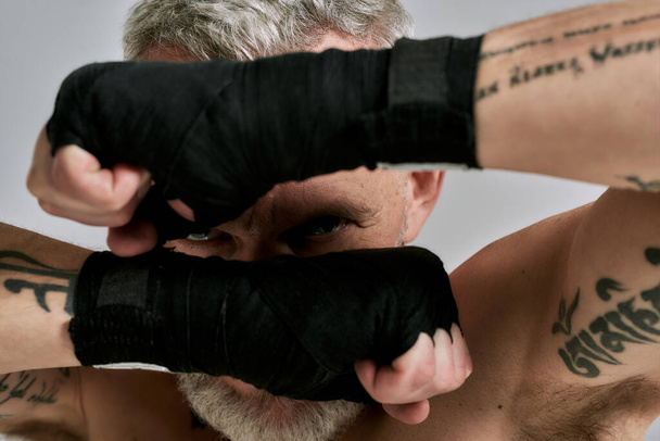 Close up of middle aged athletic man, kickboxer hiding his face behind wrapped hands in studio over grey background. Muay Thai, Boxing or Kickboxing concept - Φωτογραφία, εικόνα