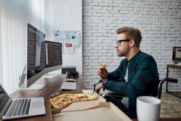 Jakie smaczne. Widok z boku kaukaski mężczyzna web developer na wózku inwalidzkim jedząc pizzę i patrząc na wiele ekranów komputerowych z kodem programu podczas siedzenia w jego miejscu pracy - Zdjęcie, obraz