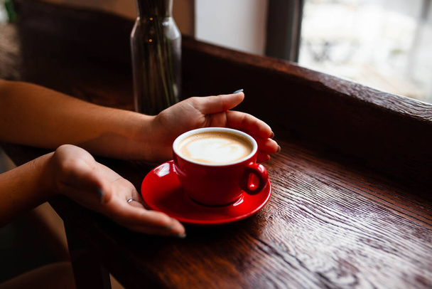 Cup of cappuccino in the hands of a young girl. Girl holding a red cup with coffee on a wooden background, hand plan and coffee. Top view. Place for text. Cappuccino with latte. - Foto, Imagen
