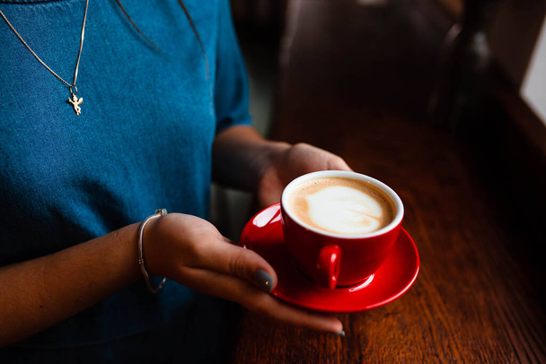Cup of cappuccino in the hands of a young girl. Girl holding a red cup with coffee on a wooden background, hand plan and coffee. Top view. Place for text. Cappuccino with latte. - 写真・画像