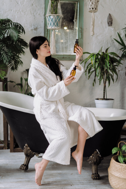 Side view of woman spending morning at bathroom, sitting on bath, choosing glass bottle with cosmetic. Eco organic care product. Vertical photo of young adult girl in bathrobe - Photo, Image