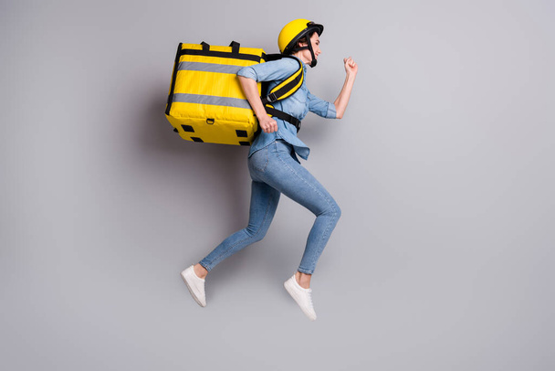 Full length profile side photo of girl courier jump run hurry deliver food home carry yellow thermal container wear denim jeans shirt shoes isolated over gray color background - Zdjęcie, obraz