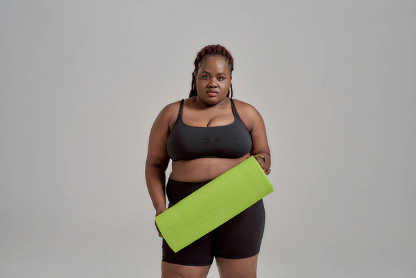 Plump, plus size african american woman in sportswear holding green yoga mat for fitness, posing in studio over grey background - Photo, image