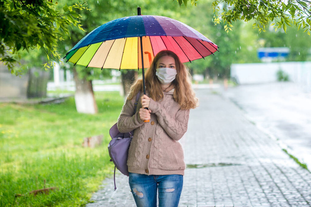 Caucasian girl in a protective mask walks under an umbrella on an empty street in spring rain. Safety and social distance during a coronavirus pandemic. New normal, the implications of quarantine - Fotografie, Obrázek