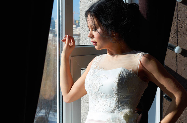 panna młoda stoi przy oknie i patrzy na suknię ślubną camera.sun - Zdjęcie, obraz