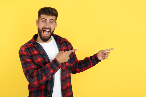 Delighted bearded hipster young man in plaid shirt point fingers away over yellow background. Look in camera. - Foto, Bild