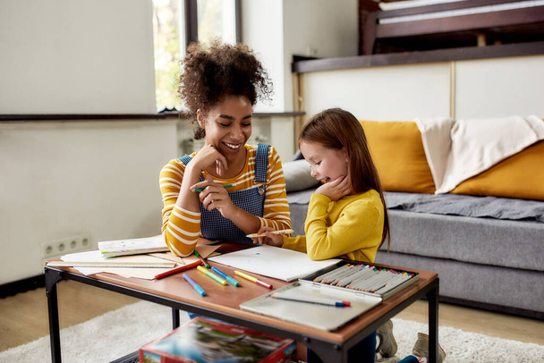 Experience childcare as it should be. Caucasian little girl spending time with african american baby sitter. They are drawing, learning how to draw, sitting on the floor - Foto, Imagem
