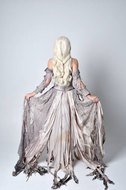 full length portrait of blonde girl wearing torn and ripped old bridal gown. standing pose with back to the camera  against a studio background. - Photo, Image