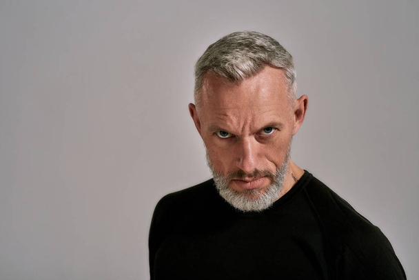 Close up of middle aged man in black t shirt looking at camera while posing in studio over grey background - Foto, afbeelding