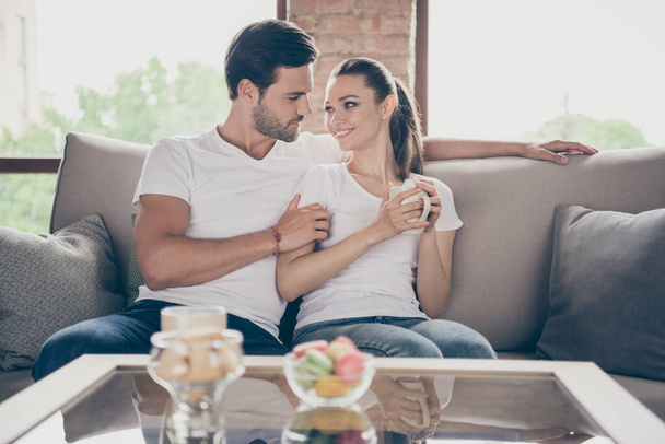 Photo of pretty charming lady handsome guy couple relaxing sit comfy couch look eyes stay home good mood quarantine hugging spend time together drink hot tea mug living room indoors - Foto, Bild