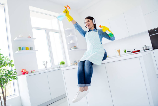 Low angle view photo of cheerful positive funky woman professional cleanup worker sit table hold sprayer spray disinfect kitchen listen headset music song sound melody in house indoors - Foto, immagini