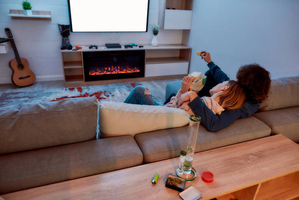 Back view of young couple watching TV together, eating snacks after smoking marijuana from a bong or glass water pipe. They relaxing on the couch in the evening at home - Zdjęcie, obraz