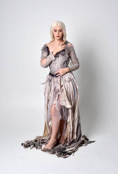 full length portrait of blonde girl wearing long torn old wedding dress.  standing pose with back to the camera on a studio background. - Photo, Image