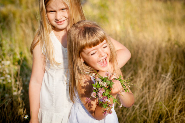 Two little friends hug and laugh merrily in the meadow, with a bouquet of wild flowers - Foto, imagen