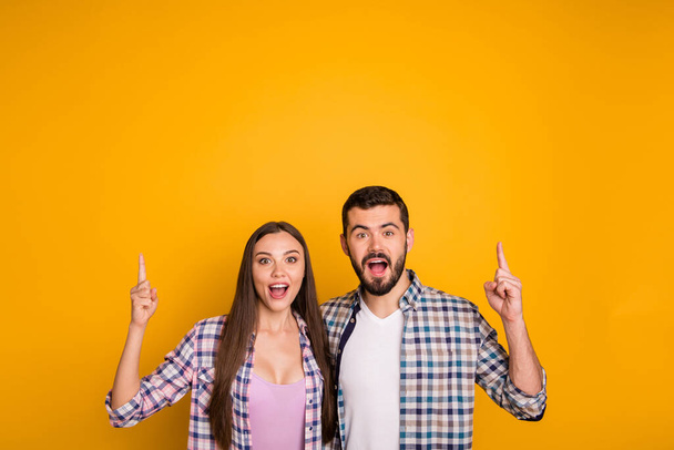 Photo charming lady handsome guy couple hugging showing novelty product directing fingers up empty space open mouth cool offer wear casual plaid shirts isolated yellow color background - 写真・画像