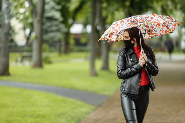 Девушка в маске идет по улице. Девушка в защитной маске гуляет по парку с зонтиком под дождем. Коронавирусная инфекция COVID-19 - Фото, изображение