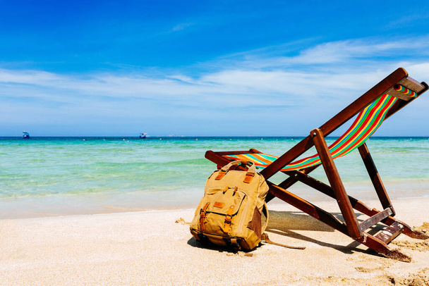 A deck chair on the sandy shore of a tropical beach overlooking the open sea. a stylish brown backpack standing nearby. A place for the traveler, freelancer. Blue sky with white clouds and bruise sea in the background - 写真・画像