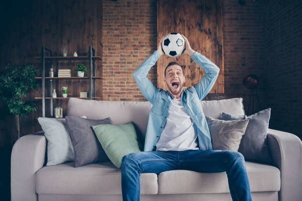 Photo of domestic handsome guy relaxing stay home quarantine time watch football match champion league europe raise leather ball yelling sit cozy sofa modern interior living room indoors - Foto, Bild