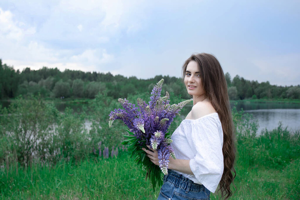 Beautiful young woman with dark hair in nature. The flowers are lupines. Summer background. - Photo, image