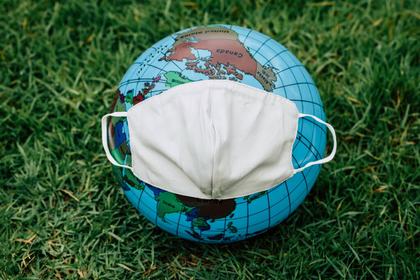 Küre küre modeli kuklası. Cerrahi maske tak. Yeşil bir çimenliğe yerleştirilmiş. Dünya topunun kavramsal resmi. Covid-19 Coronavirus 'a karşı filtre maske takıyor. (Eski tarz) - Fotoğraf, Görsel
