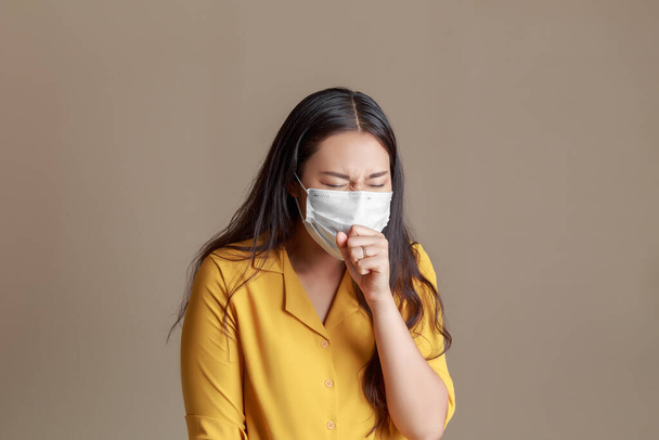 Sarı gömlekli çekici bir Asyalı kadının boğaz alerjisi var ve yüz maskesi takmış öksürüyor, hapşırıyor ve öksürüyor..  - Fotoğraf, Görsel