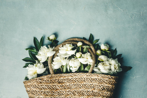Flat-lay of Beautiful peony flowers in straw bag over blue rustic background - Photo, image