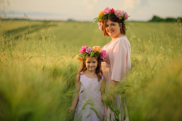 緑の小麦畑にピンクのドレスの母と娘。頭には花冠があり. - 写真・画像