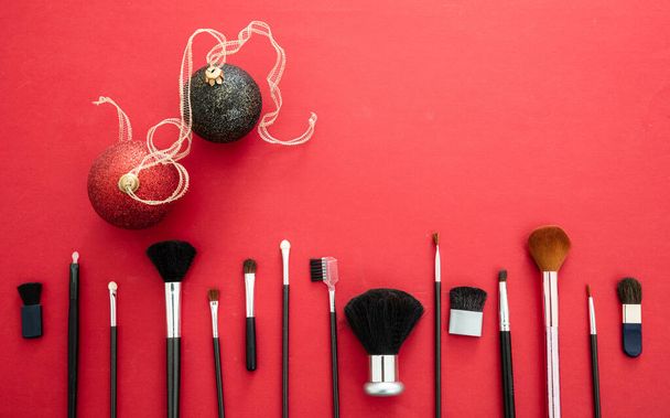 Schminken Weihnachtsfeier Konzept. Make-up Pinsel Set und Weihnachtskugeln vor rotem Hintergrund, Draufsicht. Professionelle Werkzeuge für Make-up - Foto, Bild
