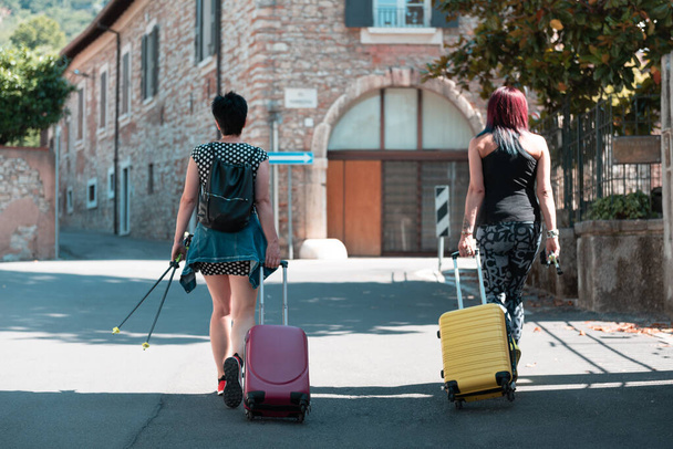 Handkoffer. Zwei Frauen gehen und wälzen ihre Rollkoffer auf dem Bürgersteig. Das Gepäck befindet sich auf der Straße der Stadt und ist reisefertig. Rückansicht von zwei Mädchen unterwegs. - Foto, Bild