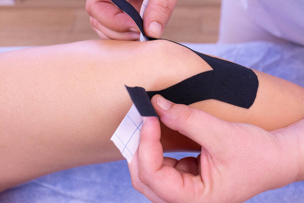 Mains d'une physiothérapeute féminine méconnaissable plaçant du ruban adhésif noir sur le genou d'une femme - Photo, image