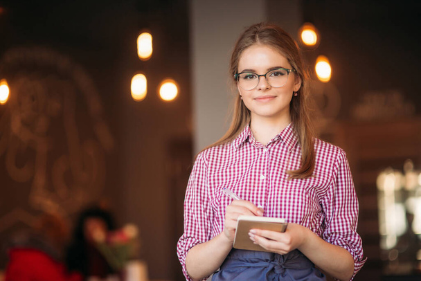 Portrait of a beautiful friendly waitress in glasses with notepapers and pen ready to take your order - Фото, зображення