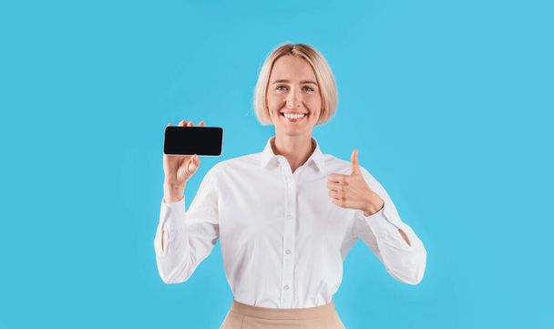 Деловая женщина держит мобильный телефон с пустым пространством для вашего дизайна и показывает большой палец вверх жеста, синий фон
 - Фото, изображение