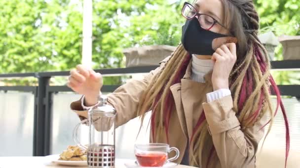 Jeune fille étudiante avec de longues dreadlocks et masque visage textil noir se démasque et commence à boire un thé aux fruits et à manger croissant à la banane dans un café après avoir ouvert des cafés de rue - Séquence, vidéo