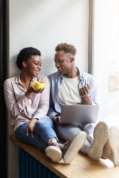 Happy black couple with laptop and coffee sitting on windowsill at cafe - Photo, Image