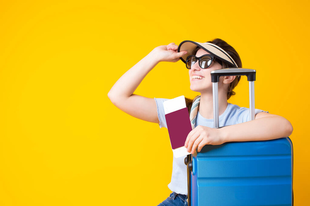 Happy smiling young brunette woman wearing summer clothes,sunglasses,straw beach cap visor,holding passport with tickets,sitting with blue suitcase and looking up away isolated over yellow background. - Photo, Image