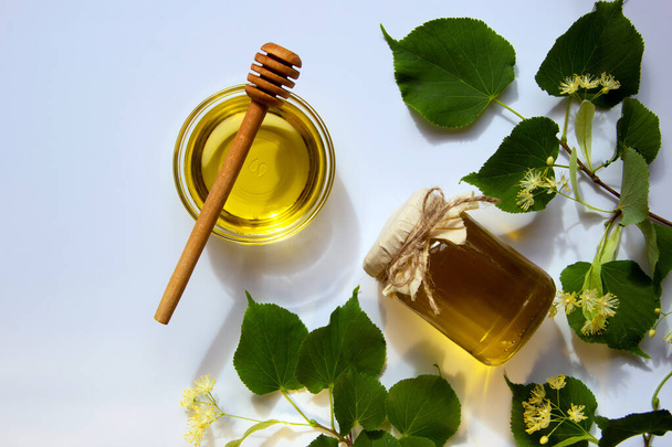 Glass jar with honey, a round glass plate, a round wooden stick for honey, a branch of a blossoming linden. Vitamins concept, natural sweets. Flat lay, copy space. - Photo, Image