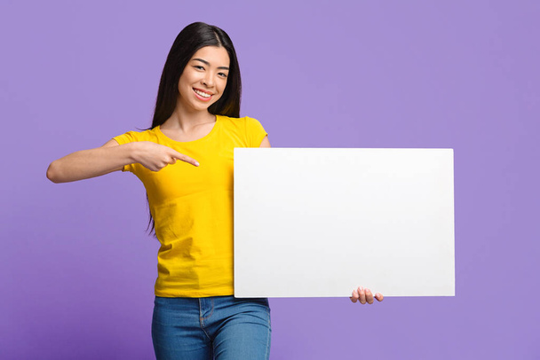 Lugar para o seu anúncio. Sorrindo menina asiática apontando para branco em branco Placard
 - Foto, Imagem